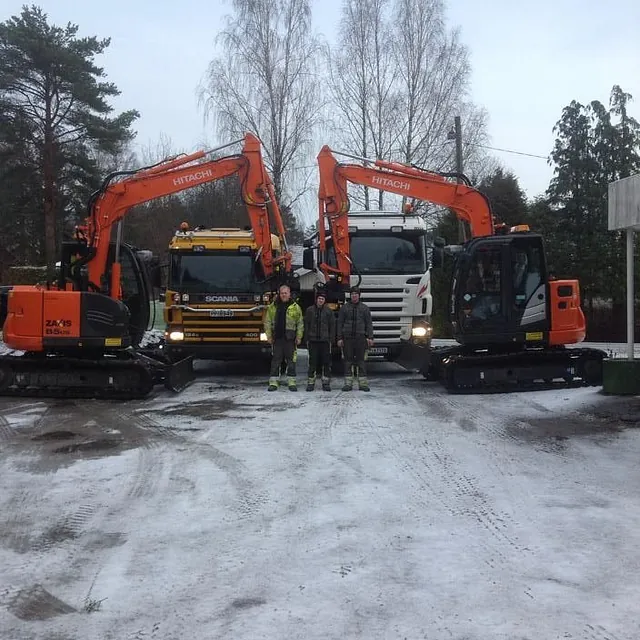 To Hitachi gravemaskiner og lastebiler parkert på snødekt bakke, med tre personer foran.