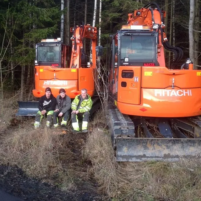 Tre personer sitter foran to oransje Hitachi gravemaskiner parkert ved en skogkant.